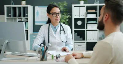Médica de jaleco e estetoscópio conversando com um paciente do outro lado da mesa do consultório