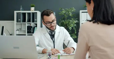 Médico sentado à mesa do consultório conversando com uma paciente enquanto faz anotações