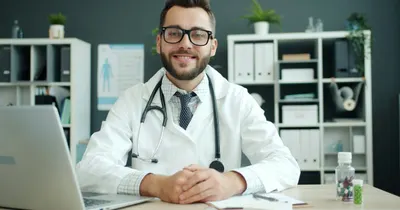 Médico jovem de jaleco e estetoscópio sorrindo, sentado à mesa do consultório com um notebook