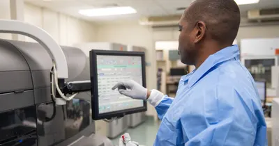 Profissional de laboratório de avental azul e luvas operando a tela de um analisador bioquímico