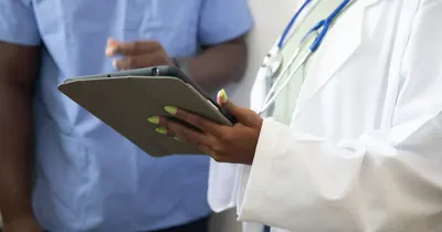 Mãos de uma médica de jaleco segurando um tablet ao lado de um colega de uniforme azul