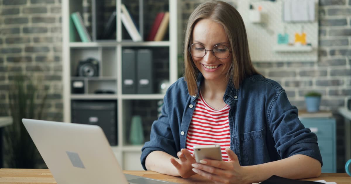 Mulher sentada à mesa de trabalho lendo uma mensagem no celular
