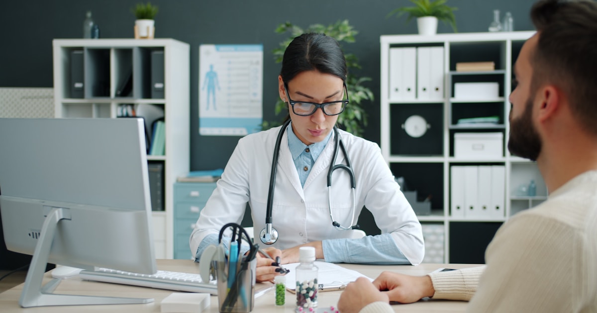 Médica conversando com um paciente durante uma consulta presencial, com computador e anotações sobre a mesa