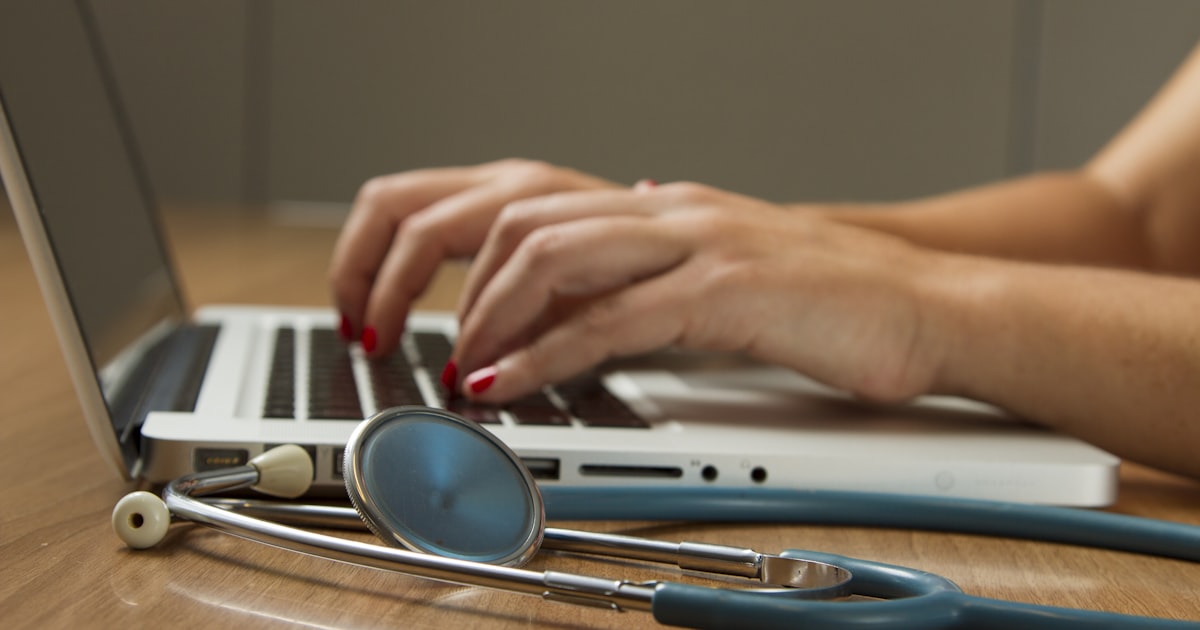 Mãos digitando em um notebook, com estetoscópio ao lado sobre a mesa do consultório